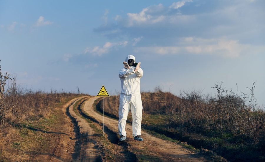 Scientist in radiation suit and gas mask showing that entrance on territory is forbidden, standing near yellow triangle with skull and crossbones sign and warning about poisonous substances and danger